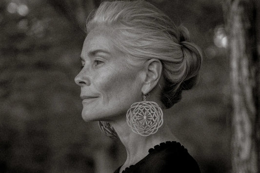 Black and white profile portrait of a woman in her 40s or 50s with silver hair styled in an updo, wearing ornate mandala-style drop earrings, photographed outdoors with a soft blurred natural background.
