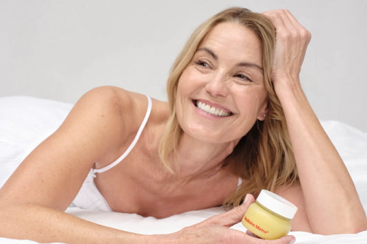 Smiling woman lying in bed holding a Medicine Mama vulva balm jar, a gentle moisturizer for perimenopause vaginal itching and vulvar dryness relief.