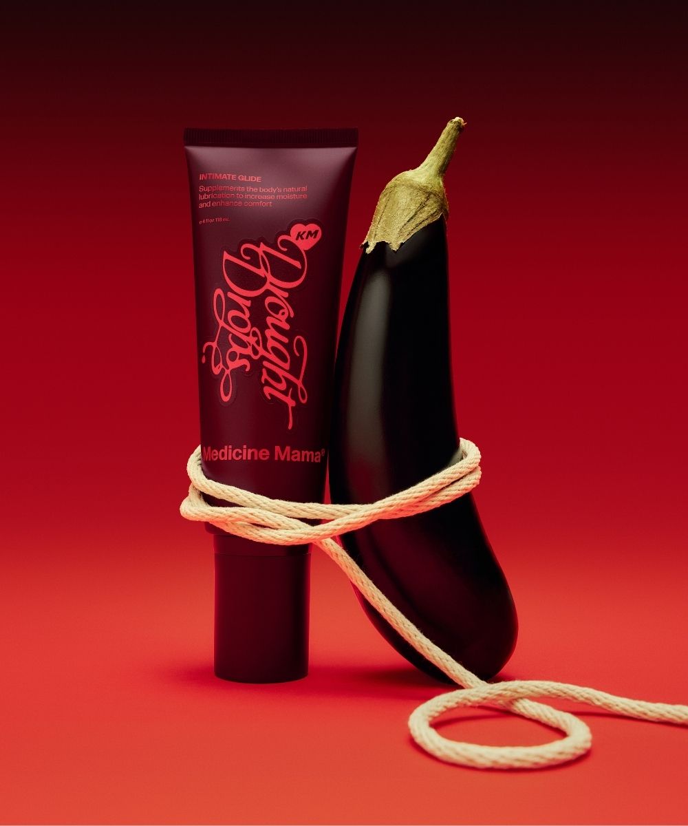 A tube labeled Drought Drops, known for its water-based personal lubricant formula, stands upright next to an eggplant, both wrapped loosely with a white rope, set against a red gradient background.