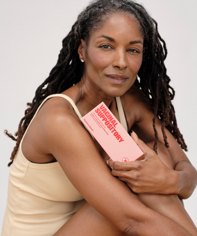 A woman with long braided hair wearing a beige tank top sits, smiling slightly, and holds a pink box labeled Vaginal Suppository against her chest.