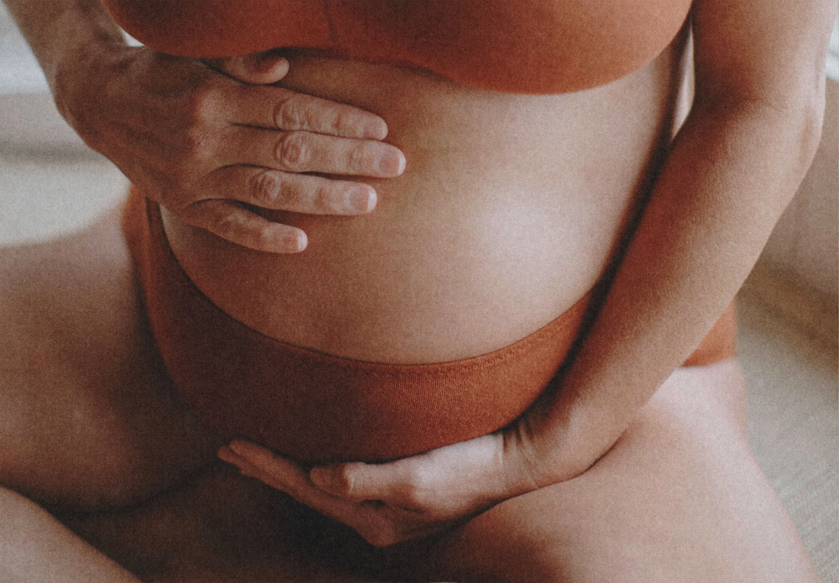 A person wearing rust-colored underwear gently cradles their pregnant belly with both hands, one above and one below, highlighting a calm, intimate moment.