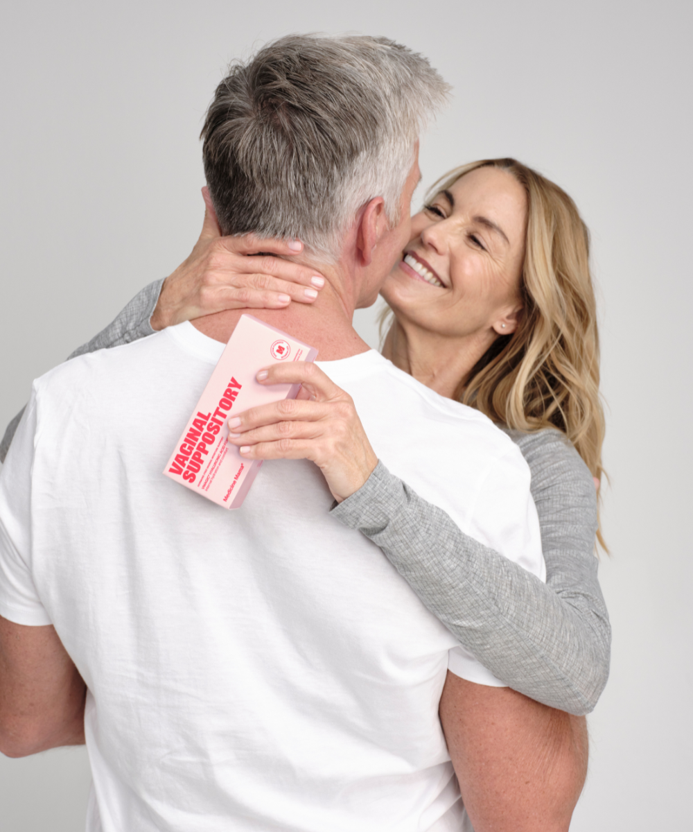 A smiling woman embraces a man from behind, holding a box labeled VMAGIC® Intimate Moisturizer Duo—a hormone-free solution for vaginal dryness. They look happy and affectionate, dressed casually against a plain background.