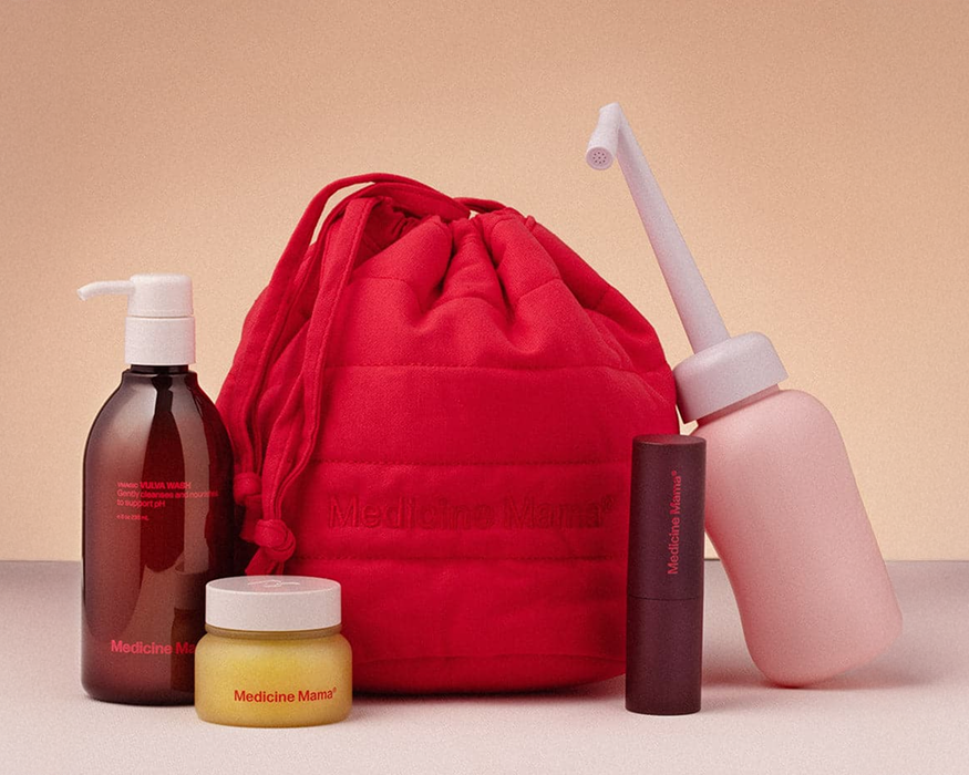 A red drawstring bag with “Medicine Mama” branding is surrounded by skincare products: a pump bottle, a jar, a cylindrical tube, and a pink peri bottle, all displayed on a light surface with a warm background.