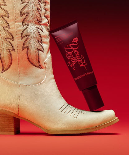 A beige cowboy boot stands next to a maroon tube labeled Drought Drops—an organic aloe lubricant that also serves as a gentle vaginal moisturizer—set against a red gradient background.