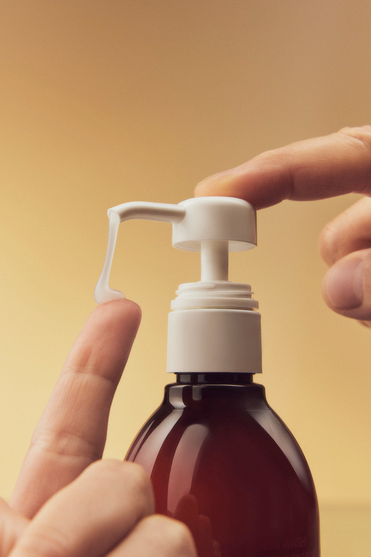 A hand presses the pump of a brown bottle, dispensing white lotion onto the index finger of another hand, against a warm beige background.