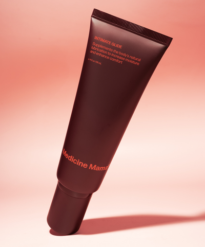 A maroon tube labeled “Intimate Glide - Preorder Ships 9/18” is displayed upright against a pink background. The label notes that it supplements moisture and enhances comfort, emphasizing its gentle, water-based formula as an organic personal lubricant and vaginal moisturizer.