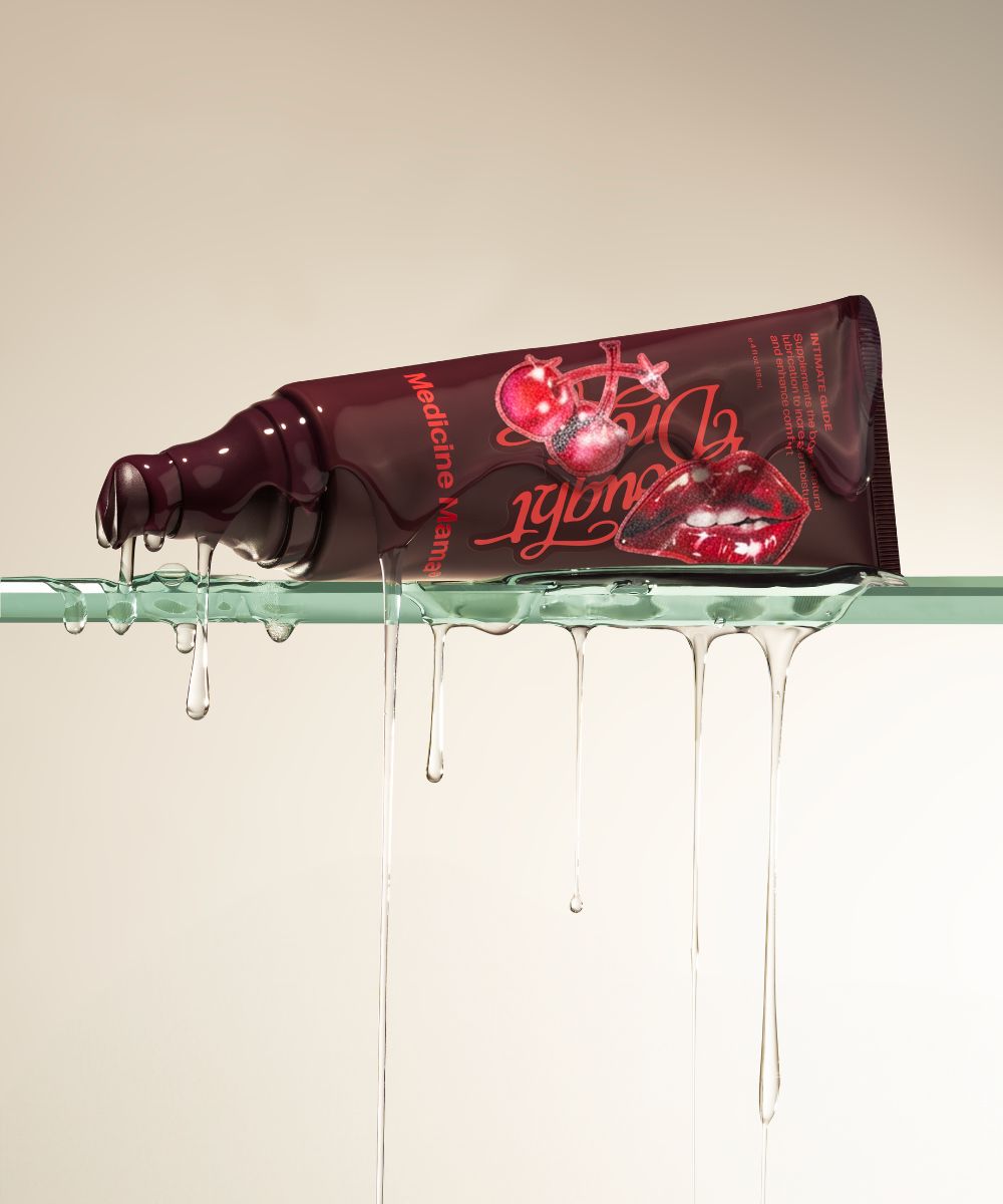 A tube of Drought Drops, an organic cherry-flavored personal lubricant, rests on a glass surface with clear, viscous liquid dripping down. The packaging showcases cherry illustrations and bold red text against a light, neutral background.