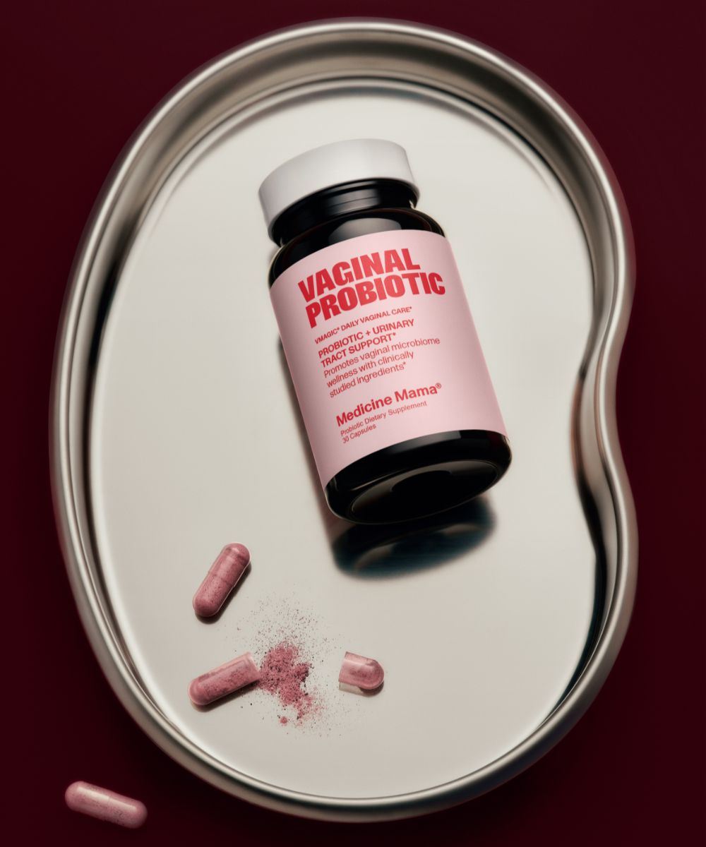 A bottle labeled VMAGIC® Vaginal Probiotic rests on a kidney-shaped metal tray beside three pink capsules, one open and spilling powder. Against a rich dark red background, the scene suggests support for urinary tract health and feminine wellness.