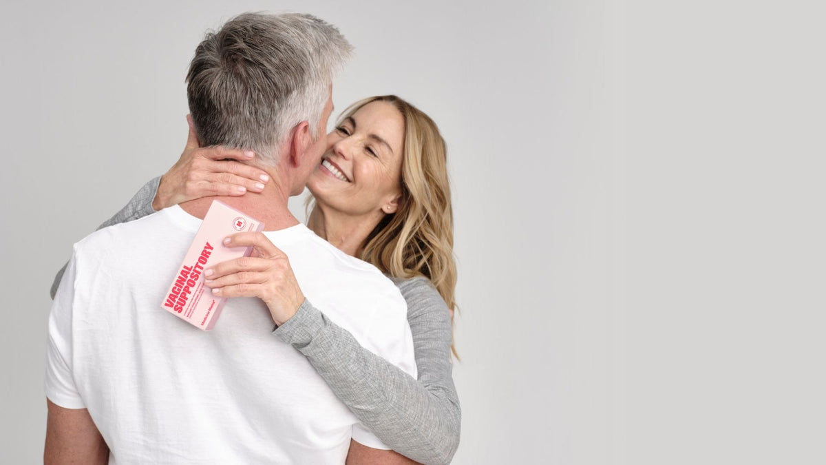 A smiling woman embraces a man, holding a box labeled Vaginal Health Story behind his back. Both appear happy and relaxed against a plain background.