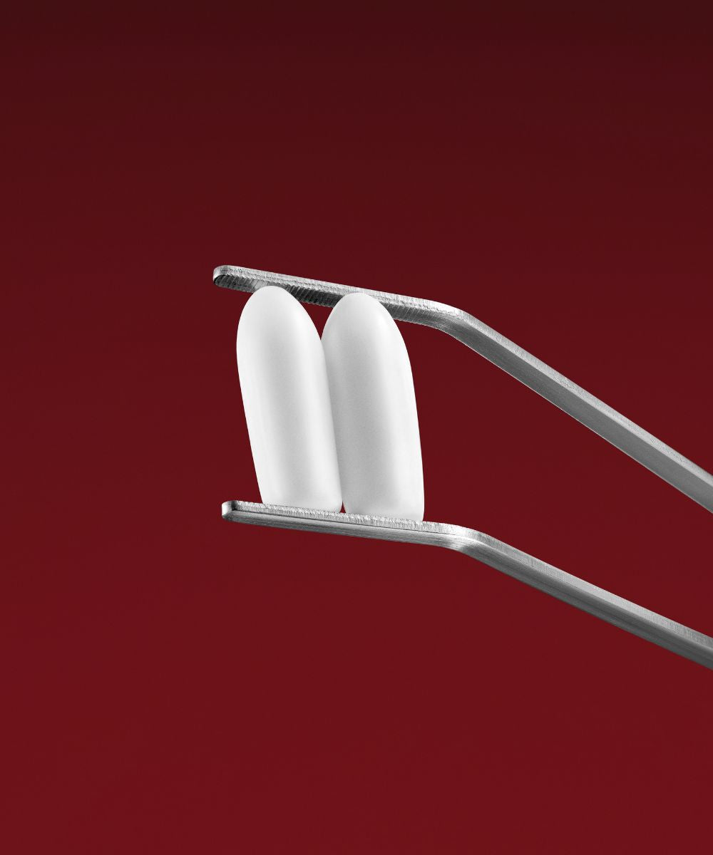 A pair of metal tweezers holds two white, oval-shaped VMAGIC® Vaginal Suppositories, designed to relieve vaginal dryness, set against a solid red background.