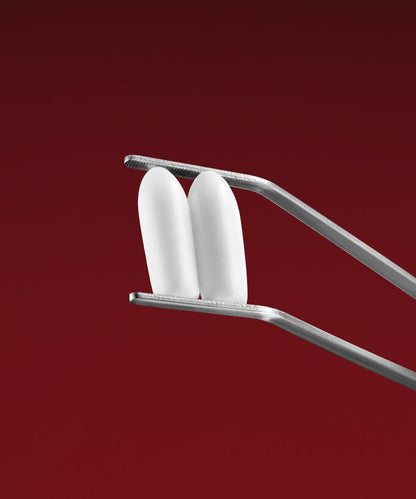 A pair of metal tweezers holds two white, oval-shaped VMAGIC® Vaginal Suppositories, designed to relieve vaginal dryness, set against a solid red background.