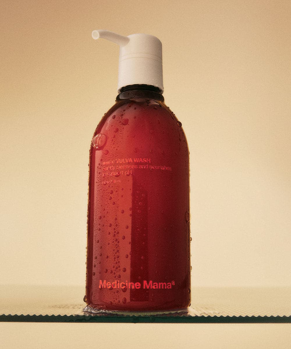 A red bottle of Medicine Mamas VIVA WASH with a white pump sits on a glass surface, covered in water droplets, against a beige background.