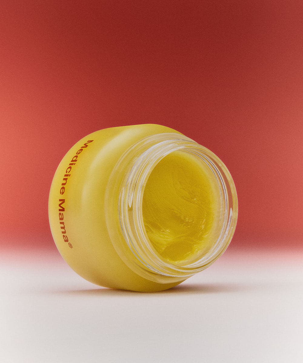A small yellow jar of cream lies on its side, open with visible yellow balm inside, against a red and white gradient background.