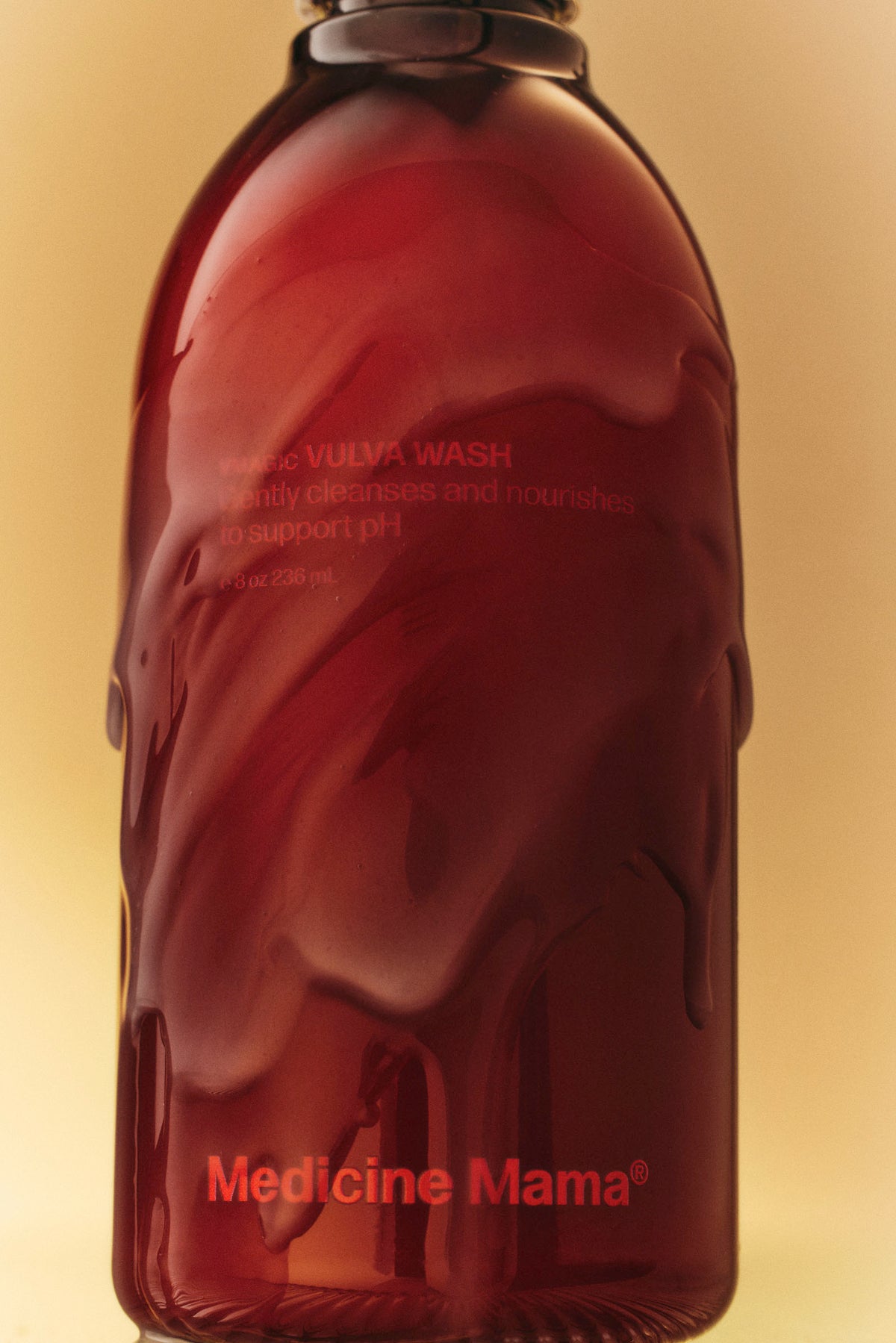 A red bottle labeled Medicine Mama with an opaque, glossy texture, and text that reads, VULVA WASH—gently cleanses and nourishes to support pH. The background is a soft yellow gradient.