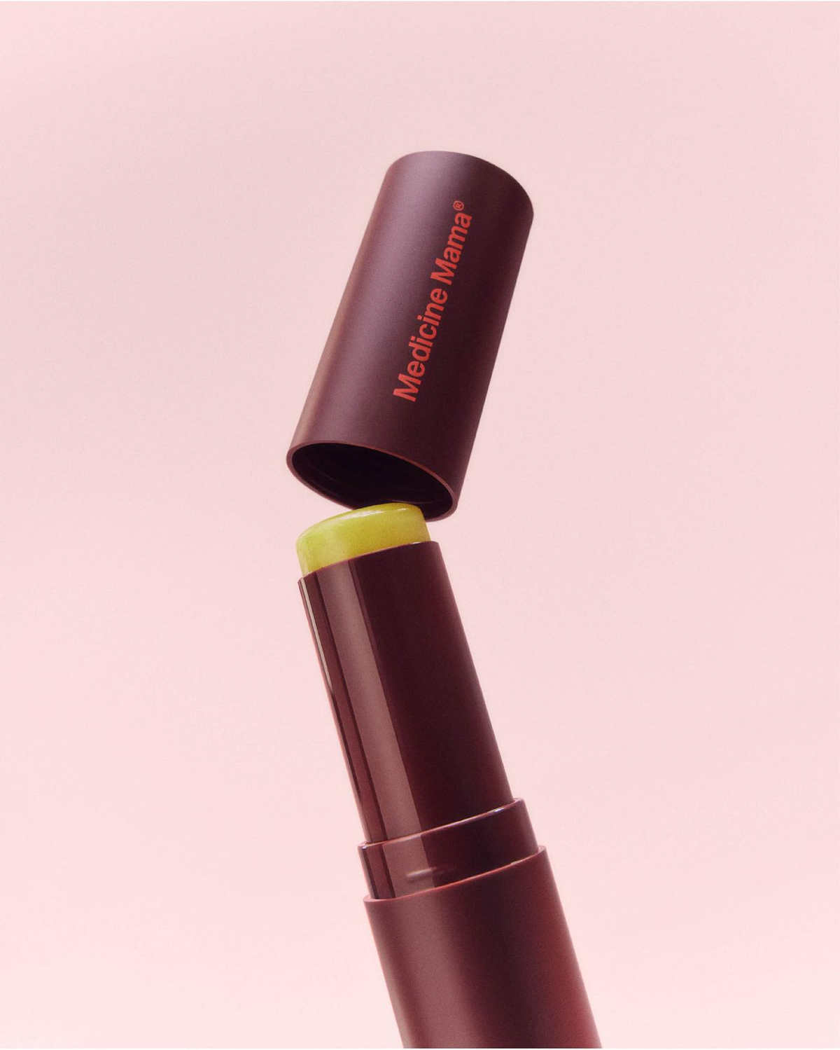 A brown lipstick-style tube with a yellow balm inside and a cap labeled Medicine Mama against a soft pink background.