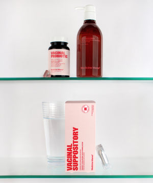 A glass of water, the VMAGIC® Vaginal + Urinary Care Kit—including a box of hyaluronic acid vaginal suppositories with a capsule beside it, a bottle labeled Vaginal Probiotic, and a red pump bottle—are arranged on two glass shelves against a white background.