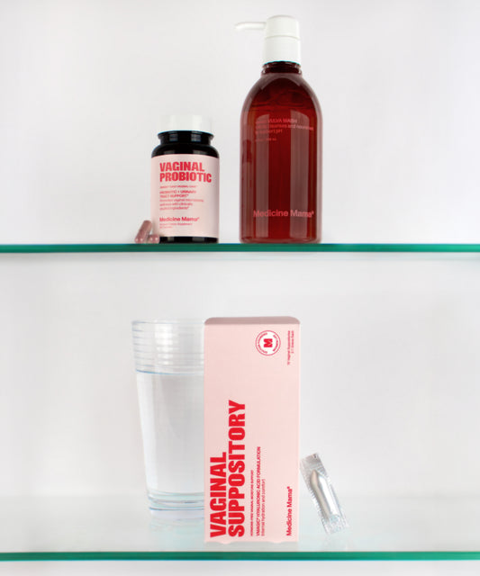 A glass of water, the VMAGIC® Vaginal + Urinary Care Kit—including a box of hyaluronic acid vaginal suppositories with a capsule beside it, a bottle labeled Vaginal Probiotic, and a red pump bottle—are arranged on two glass shelves against a white background.